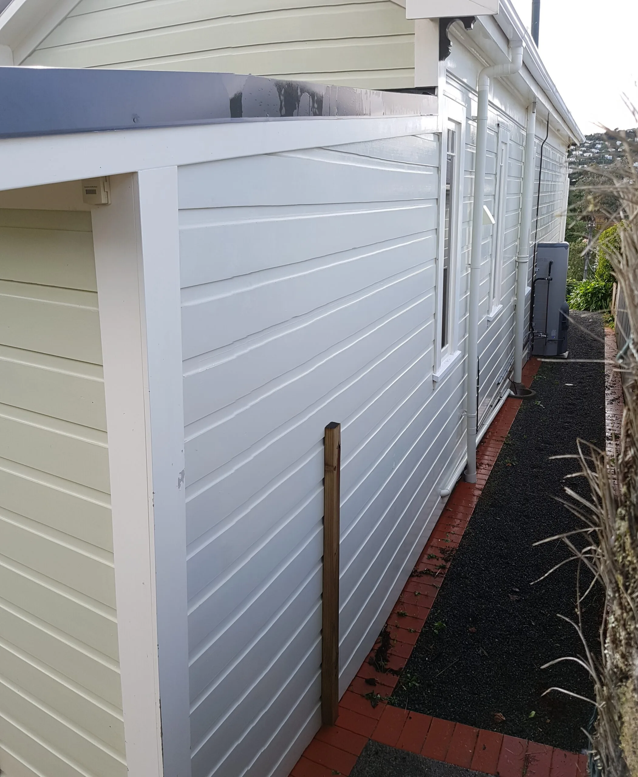 20180525_105603-scaled Side view of a white wooden building with a sloped roof and guttering, featuring neatly aligned grey and white wooden panels. a red brick path and green plants contrast with the building’s exterior.